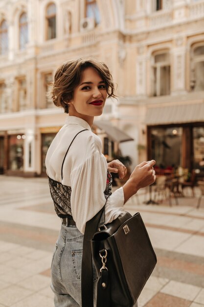 Schöne Frau mit kurzer Frisur in Jeans, die schwarze Handtasche in der Stadt halten. Wunderbare Frau im Hemd mit dunkler Spitze, die an Straße lächelt.