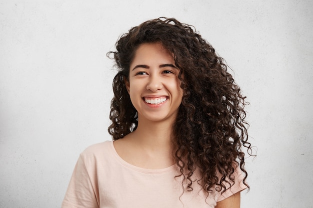 Schöne Frau mit lockigem buschigem Haar, gemischter Nationalität, lässig gekleidet, breit lächelnd, zeigt weiße perfekte Zähne