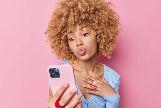 Schöne Frau mit lockigem, buschigem Haar hält die Lippen gefaltet, macht Selfie-Posen auf dem Smartphone, hat ein Online-Gespräch mit ihrem Freund, drückt ihre Liebe aus, weas lässiger blauer Pullover und Schmuck isoliert auf rosa Wand