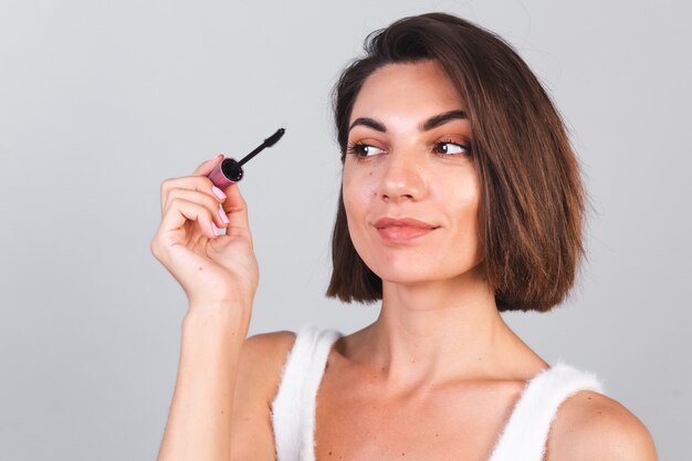 Schöne Frau mit Make-up halten schwarze Wimperntuschebürste auf grauer Wand, Schönheitskonzept