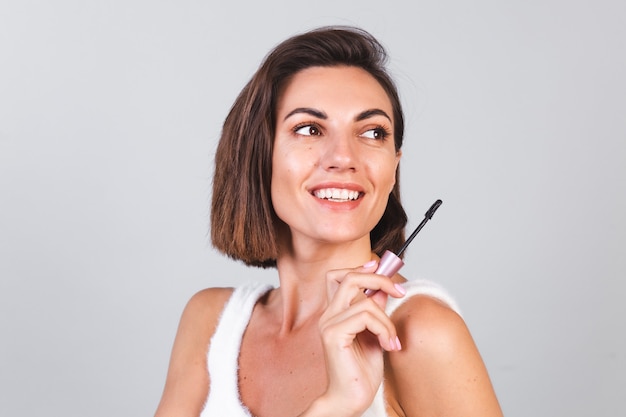 Schöne Frau mit Make-up halten schwarze Wimperntuschebürste auf grauer Wand, Schönheitskonzept