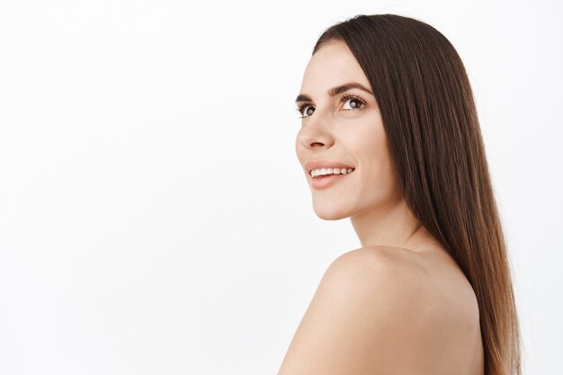 Schöne Frau mit perfekt sauberem, hydratisiertem, glänzendem Gesicht und langem Naturhaar, brünettes weibliches Modell, das das Kopienraumlogo der Hautpflege-Haarpflegekosmetik beiseite schaut und nackte Schultern steht