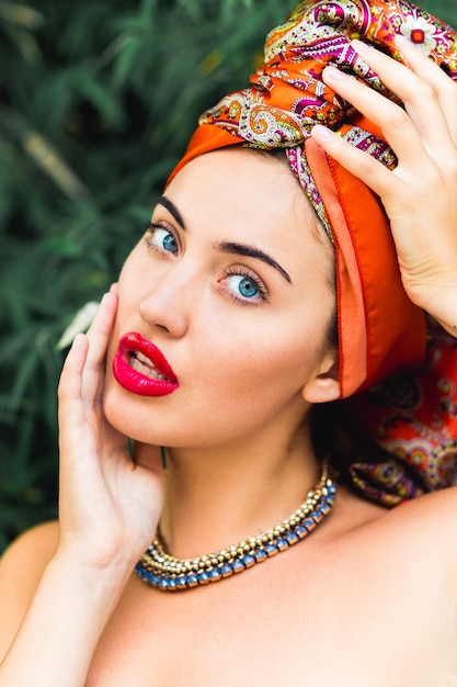 schöne Frau mit perfektem Make-up und orangefarbenem Kopftuch, roten großen Lippen, blauen Augen, Händen auf dem Kopf