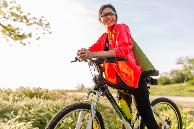 Schöne Frau mit schmaler Passform, die morgens im Park Sport auf dem Fahrrad mit Yogamatte im bunten Fitness-Outfit macht, die Natur erforscht, glücklichen gesunden Lebensstil lächelnd