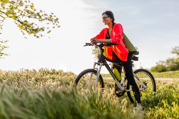 Schöne Frau mit schmaler Passform, die morgens im Park Sport auf dem Fahrrad mit Yogamatte im bunten Fitness-Outfit macht, die Natur erforscht, glücklichen gesunden Lebensstil lächelnd