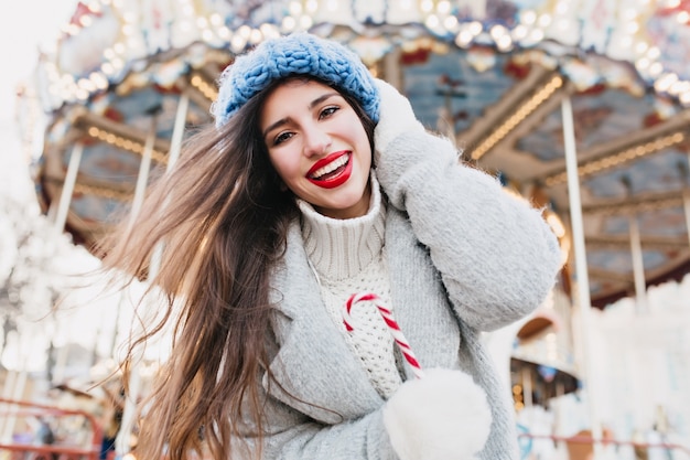 Kostenloses Foto schöne frau mit süßer zuckerstange, die nahe karussell in weihnachten aufwirft. foto im freien des glücklichen dunkelhaarigen mädchens mit lutscher, das im vergnügungspark im winter entspannt.