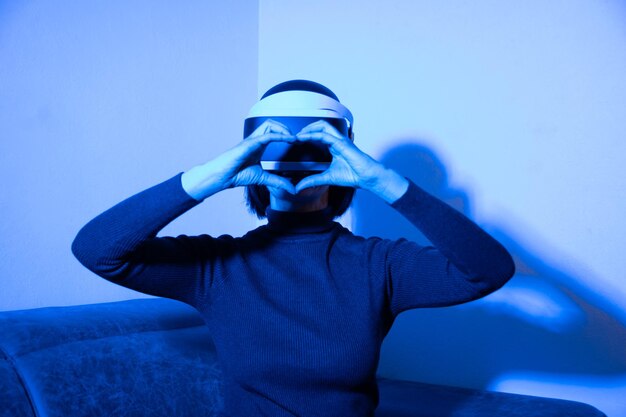 Schöne frau mit vr-headset zeigt herzform mit händen zu hause in blauem licht. das konzept der virtuellen beziehungen. virtuelles liebeskonzept.