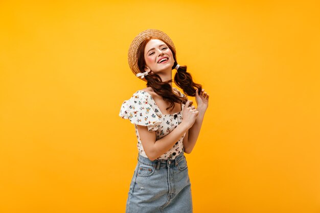 Schöne Frau mit zwei Pferdeschwänzen lächelt kokett. Frau im Hut, im Sommeroberteil und im Jeansrock, die auf orange Hintergrund aufwerfen.