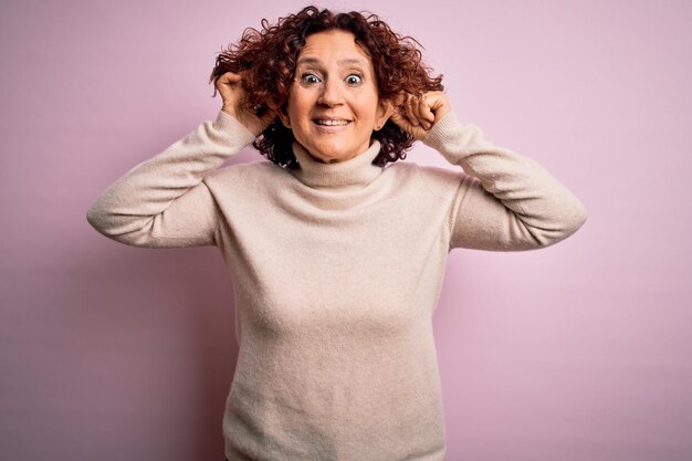 Schöne Frau mittleren Alters mit lockigem Haar trägt lässigen Rollkragenpullover vor rosa Hintergrund und zieht lächelnd die Ohren mit den Fingern. Lustige Geste. Problem beim Vorsprechen