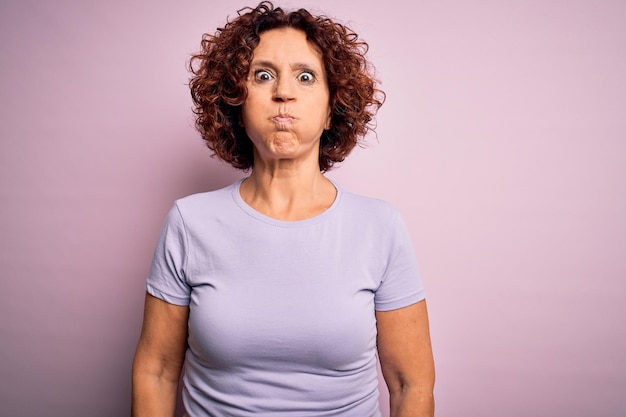 Schöne Frau mittleren Alters mit lockigem Haar trägt lässiges T-Shirt vor isoliertem rosa Hintergrund, aufgeblasene Wangen mit lustigem Gesicht, Mund mit Luft aufgeblasen, verrückter Gesichtsausdruck