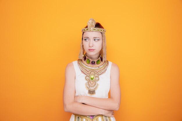 Schöne Frau wie Kleopatra im alten ägyptischen Kostüm, die beleidigt beiseite schaut, wütend auf jemanden mit verschränkten Armen auf Orange zu sein