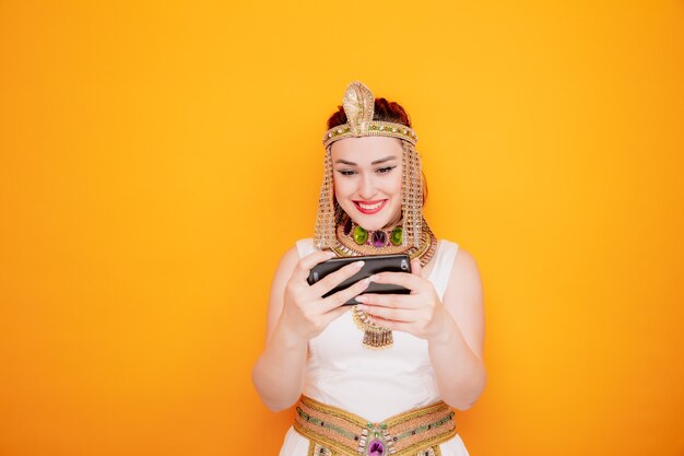 Schöne Frau wie Kleopatra im alten ägyptischen Kostüm, die Spiele mit dem Smartphone spielt, das glücklich und fröhlich auf Orange lächelt