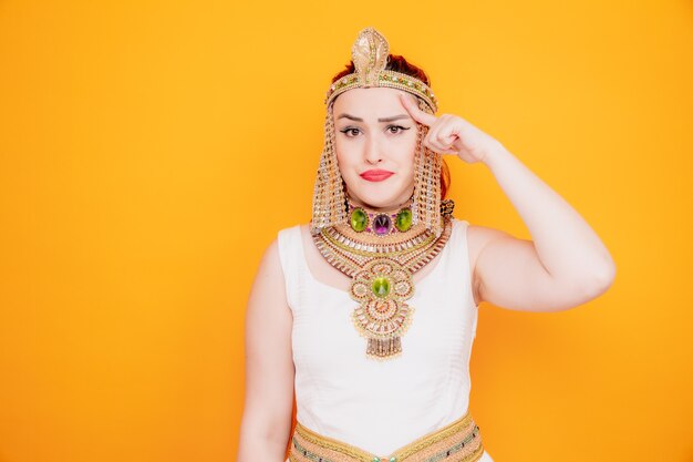 Schöne Frau wie Kleopatra in altägyptischem Kostüm verwirrt, die mit dem Zeigefinger auf ihre Schläfe zeigt und versucht, sich auf eine Aufgabe zu konzentrieren, die hart auf Orange ist