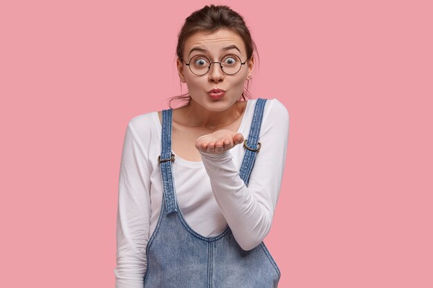 Schöne Frau zeigt Luftkusszeichen, streckt Handfläche nahe Mund, macht Grimasse, trägt runde Brille, weißen Pullover und Jean Sarafan