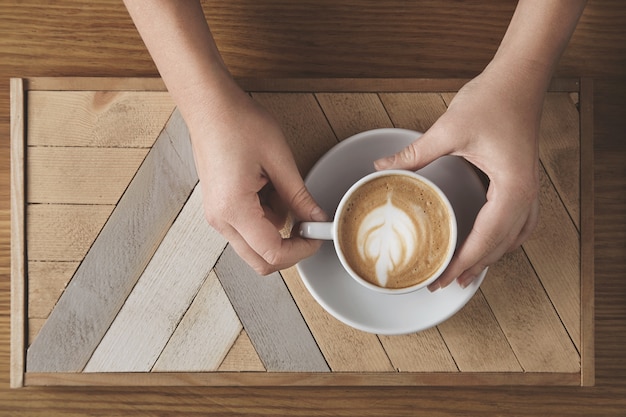 Schöne frauenhände halten keramikweiß mit cappuccino über holzteller und rustikalem tisch. milchschaum oben in baumform. draufsicht im caféladen. verkaufspräsentationskonzept.