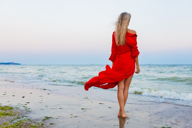 Schöne freie Frau im roten Kleid im Wind am Meeresstrand, der auf Sommer geht