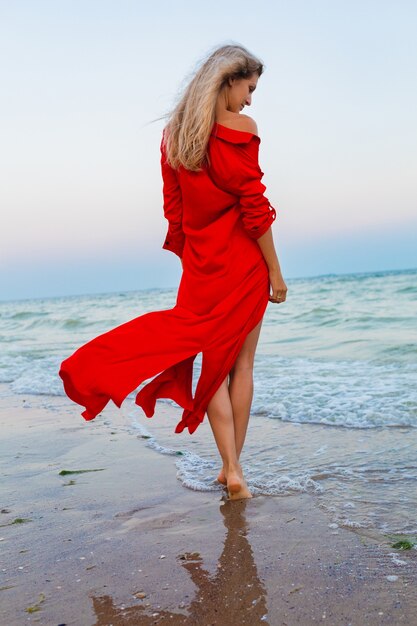 Schöne freie Frau im roten Kleid im Wind am Meeresstrand zu Fuß