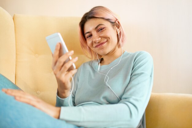 Schöne freudige junge Frau mit rosa Haaren und Piercing im Gesicht, die sich auf gelbem Sofa unter Verwendung von Handy und Ohrhörern entspannen