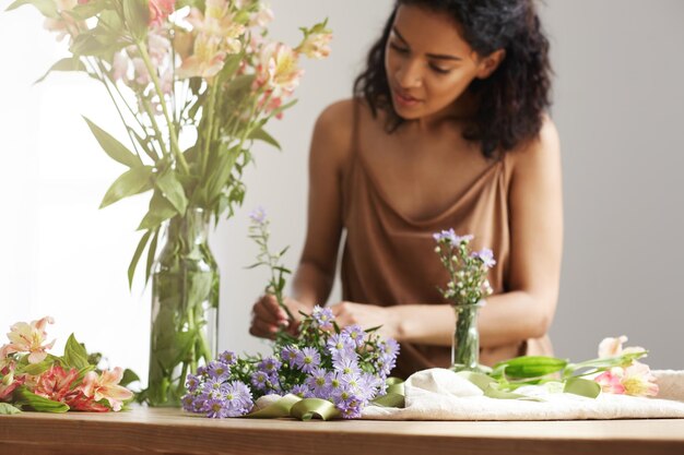 Schöne fröhliche afrikanische Mädchenfloristin, die lächelnd Blumenstrauß im Blumenladen über weißer Wand macht Kopierraum