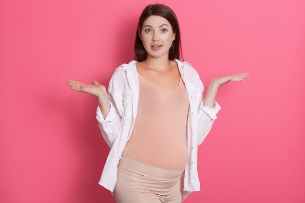 Schöne fröhliche europäische schwangere Frau im bequemen Outfit, das Hände mit überraschtem Ausdruck ausbreitet