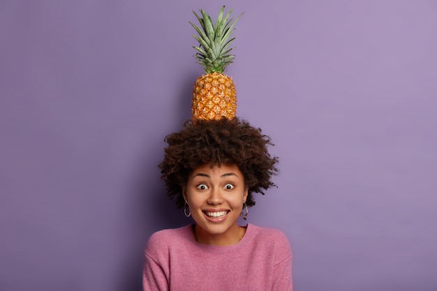 Kostenloses Foto schöne fröhliche frau mit afro-haarschnitt, hält frische ananas mit grünen blättern auf dem kopf, posiert mit sommerfrucht