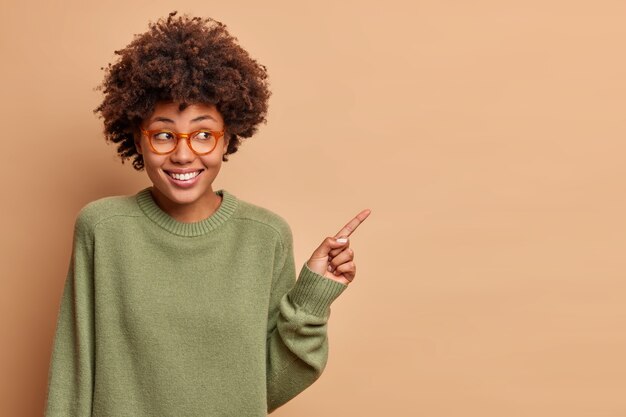 Schöne fröhliche Frau zeigt zur Seite mit Zeigefinger hat glückliches Gesicht trägt Brille lässiger Pullover zeigt Kopierraum für Ihre Werbeinhalte isoliert über braune Wand