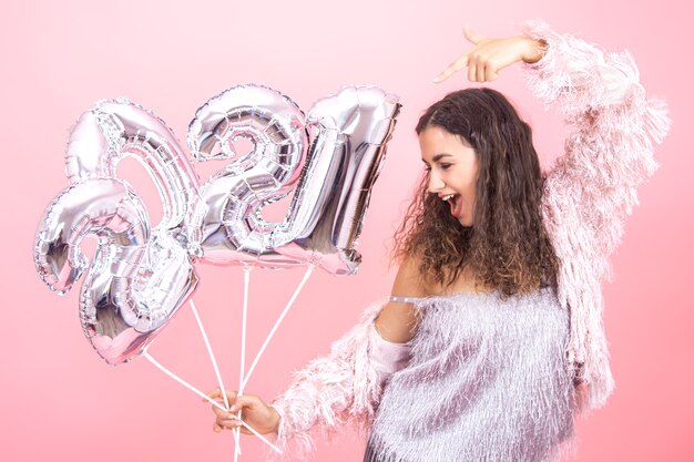 Schöne fröhliche junge Brünette mit lockigem Haar festlich gekleidet auf einem rosa Hintergrund, der souverän mit silbernen Luftballons für das Neujahrskonzept aufwirft