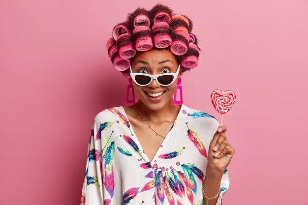 Schöne fröhliche junge Frau in trendiger Sonnenbrille, trägt Lockenwickler, macht Frisur, gekleidet in Hauskleid, hält Lutscher. Glückliche ethnische Dame wirft für Fotografen mit köstlichen Süßigkeiten auf