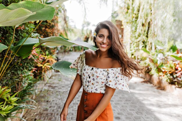 Schöne gebräunte Dame im Sommeroutfit lächelt aufrichtig gegen Wand der tropischen Pflanzen