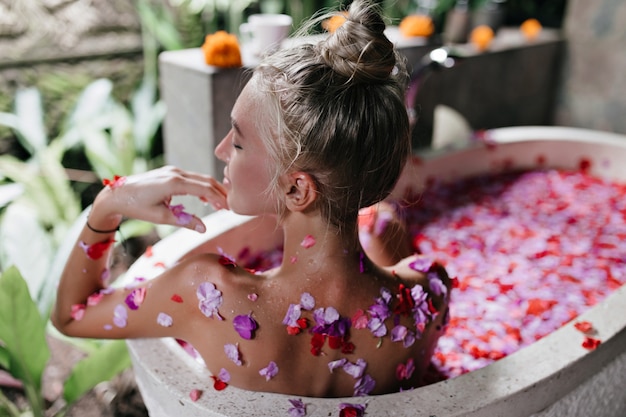 Kostenloses Foto schöne gebräunte frau, die im bad voller rosen sitzt und lächelt. innenporträt von der rückseite des niedlichen kaukasischen weiblichen modells, das während des spa nach hartem tag entspannt.