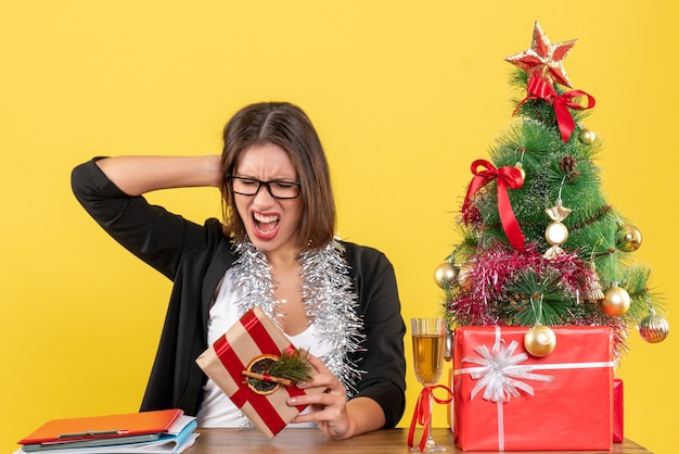 Schöne Geschäftsdame im Anzug mit Brille, die ihr Geschenk hält, das emotional an einem Tisch mit einem Weihnachtsbaum darauf im Büro sitzt