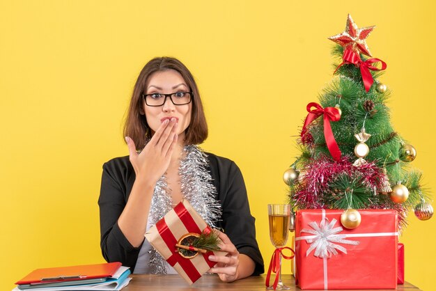 Schöne Geschäftsdame im Anzug mit Brille, die Küsse sendet und an einem Tisch mit einem Weihnachtsbaum darauf im Büro auf Gelb sitzt