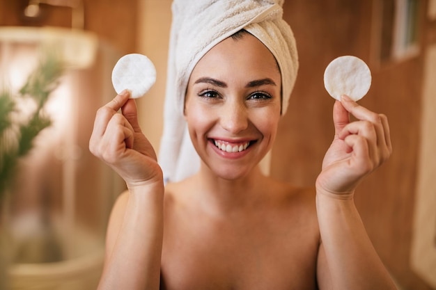 Schöne glückliche Frau, die zwei Wattepads hält, während sie im Badezimmer in die Kamera schaut