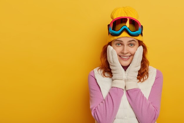 Schöne glückliche Frau hält beide Hände auf Wangen, hat zartes Lächeln, schaut mit Lächeln in die Kamera, genießt aktive Ruhe und Skitouren, gekleidet in aktiver Kleidung, isoliert auf gelber Wand. Winterzeit.