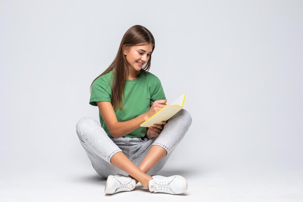Schöne glückliche junge Frau in Freizeitkleidung auf einem Boden sitzend mit gekreuzten Beinen isoliert auf weiß, ein Tagebuch haltend