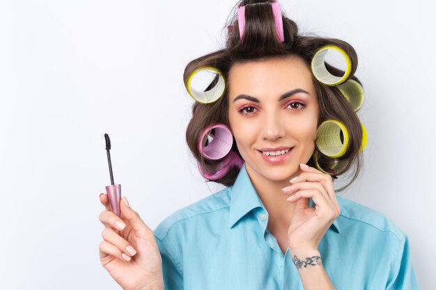 Schöne Hausfrau Eine junge, fröhliche Frau mit Haarlockenwicklern, hellrosa Make-up und Wimperntusche bereitet sich auf ein Date-Night-Dinner auf weißem Hintergrund vor