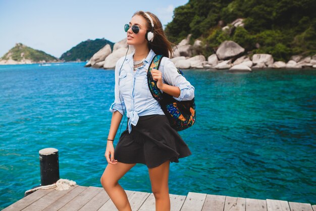 Schöne Hipster-Frau, die mit Rucksack um die Welt reist, lächelnd, glücklich, positiv, Musik in Kopfhörern hörend, blauer tropischer Ozeanhintergrund, Sonnenbrille, sexy, Sommerferien,