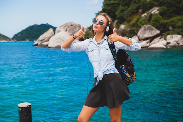 Schöne Hipster-Frau, die mit Rucksack um die Welt reist, lächelnd, glücklich, positiv, Musik in Kopfhörern hörend, blauer tropischer Ozeanhintergrund, Sonnenbrille, sexy, Sommerferien,