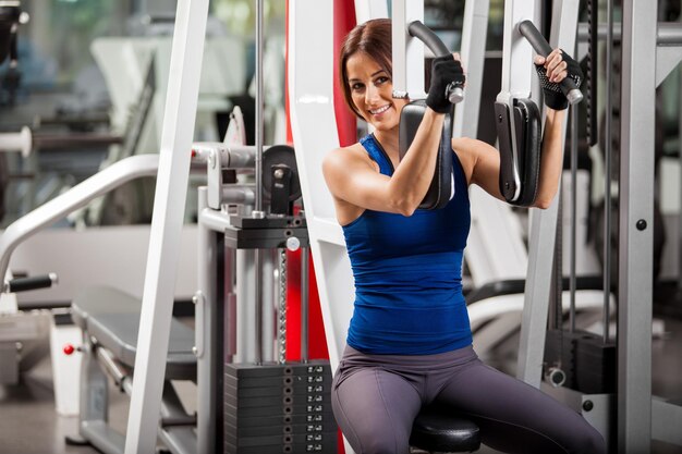 Schöne hispanische junge Frau, die in einem Simulator in einem Fitnessstudio trainiert und lächelt