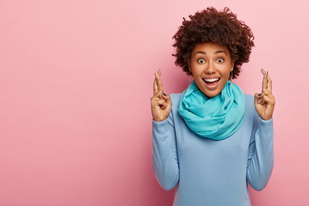 Schöne hoffnungsvolle junge afroamerikanische Frau drückt die Daumen für viel Glück, glaubt, dass Träume wahr werden, trägt blaue Freizeitkleidung, solated über rosa Studiowand.