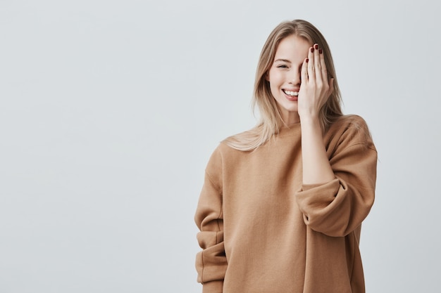 Schöne hübsche charmante junge blonde Frau im losen Pullover, der glücklich lächelt, Spaß drinnen hat und ein Auge mit der Hand schließt