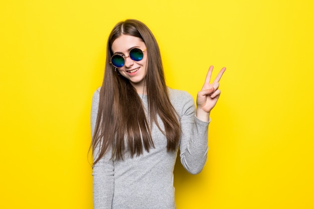 Schöne hübsche Frau, die das V-Zeichen nahe Augen macht, die elegantes Kleid tragen, das auf gelber Wand lokalisiert trägt