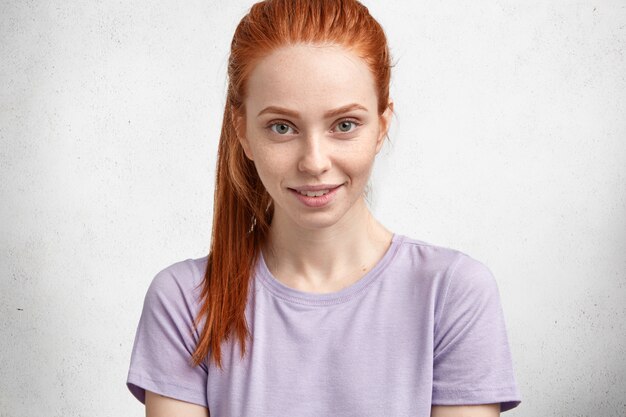 Schöne Ingwerfrau im lässigen lila T-Shirt, schaut mit erfreut erfreutem Ausdruck an der Kamera, stellt gegen weiße Betonwand auf.