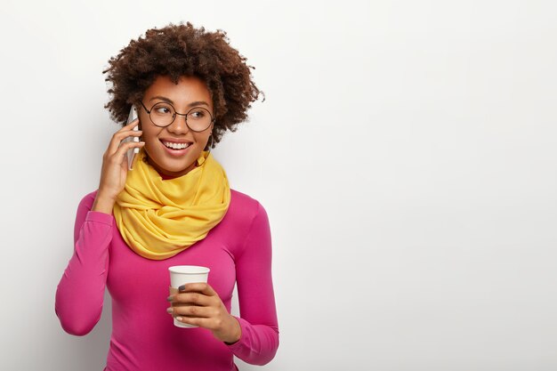 Schöne junge Afroamerikanerin hat fröhlichen Ausdruck, hat Telefongespräch, trinkt Kaffee zum Mitnehmen, fröhlichen Ausdruck, trägt transparente runde Brille, gelben Schal und rosa Rollkragenpullover