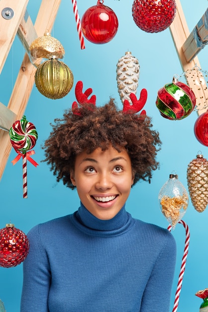 Schöne junge Afroamerikanerin mit lockigem Haar schaut über Lächeln fröhlich gekleidet in lässigem Poloneck, umgeben von Neujahrsspielzeug