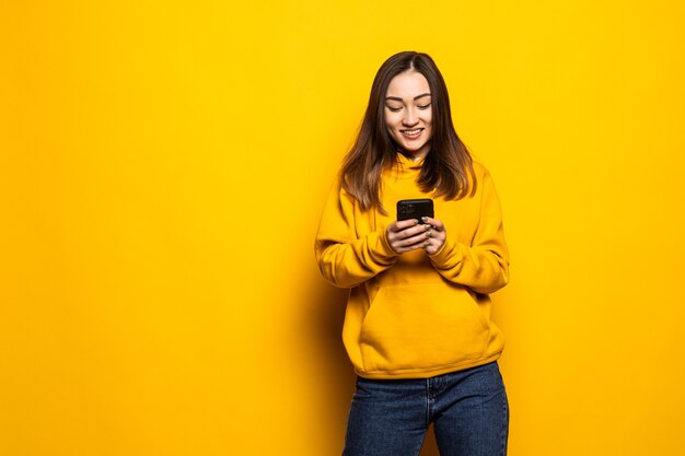 Schöne junge asiatische Frau des Porträts benutzt intelligentes Handy auf gelber Wand