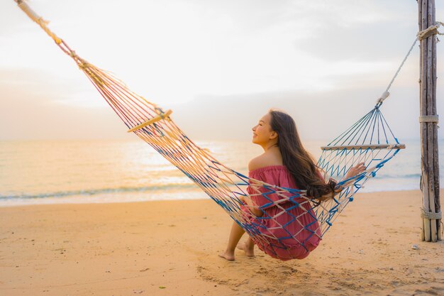 Schöne junge asiatische Frau des Porträts, die auf der Hängematte mit glücklichem neary Strandmeer und -oce des Lächelns sitzt