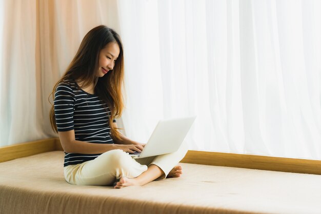 Schöne junge asiatische Frau des Porträts, die Computernotizbuch oder Laptop auf Sofa im Wohnzimmer verwendet