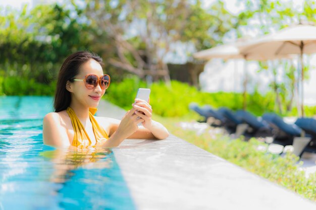 Schöne junge asiatische Frau des Porträts, die Handy oder Mobiltelefon im Swimmingpool verwendet