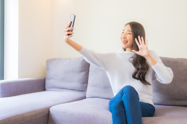 Schöne junge asiatische Frau des Porträts, die intelligentes Handy auf Sofa im Wohnzimmerinnenraum verwendet